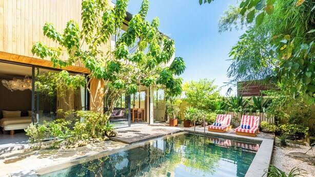 A modern outdoor pool area with a glass-enclosed lounge, surrounded by lush greenery and a wooden deck. - Hygge Villa Ubud (Ubud)