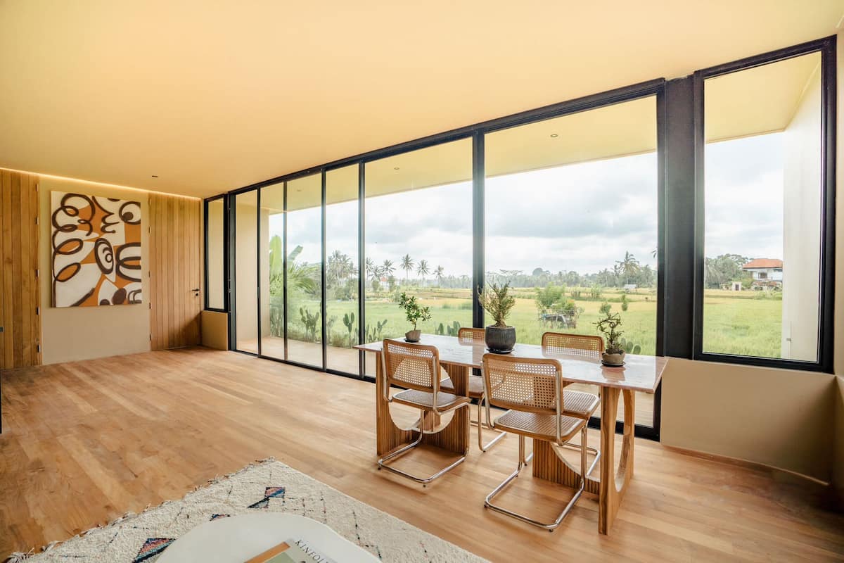 Amplia habitación con un ventanal grande, suelo de madera, una mesa de comedor con sillas y una planta en maceta.