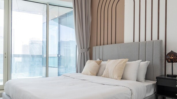 A modern bedroom with a large bed, a nightstand, and a city view through floor-to-ceiling windows. - Luxe 4bed Apt Dubai Marina JBR Pool by Belvilla (Dubai)