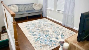 Una sala de estar con un sofá gris, un tapete estampado, una escalera de madera y un televisor.
