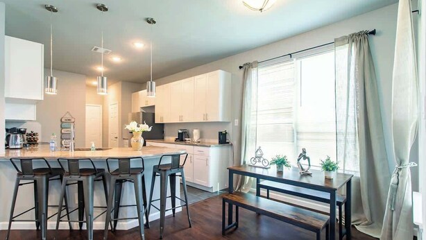 Una cocina moderna con una isla central, taburetes altos, lámparas colgantes y un comedor con banco y mesa.