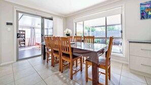 Een eetgedeelte met een grote tafel en stoelen, een keuken met witte kasten, en een schuifdeur naar een buitenruimte.