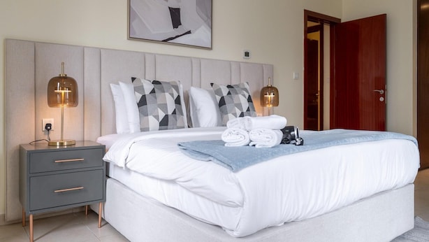 A neatly made bed with white linens, a grey bedside table with drawers, two bedside lamps, and a large abstract painting on the wall.