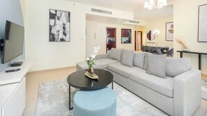 A modern living room with a grey sofa, a coffee table, and a TV on a stand. There is a chandelier above the sofa and a framed artwork on the wall.