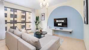 A modern living room with a sofa, a TV mounted on a blue wall, and a large window offering a view of a building.