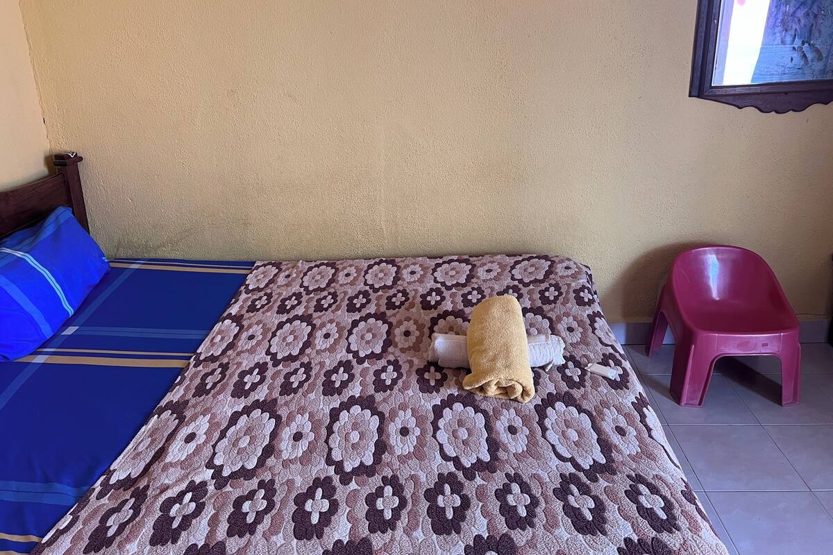A bed with a patterned bedspread, a blue and brown pillow, a rolled towel, and a pink plastic chair.