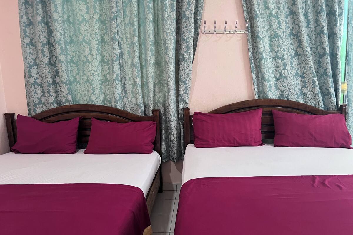 Two beds with matching purple bedding and pillows, a wooden headboard, and a window with patterned curtains.