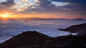 A mountainous landscape with a sunrise or sunset, cloud cover, and distant hills.