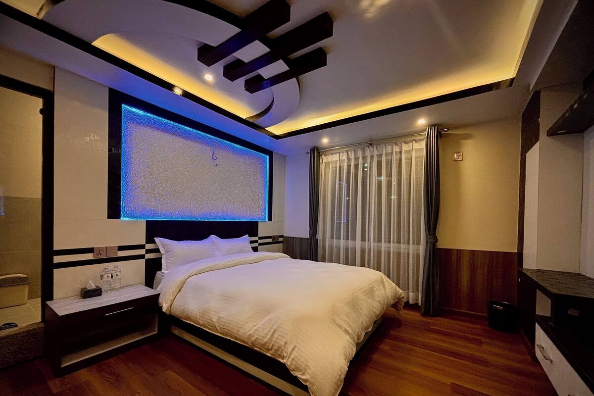 A modern bedroom with a large bed, a bedside table, a TV mounted on the wall, and a window with curtains.