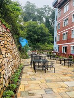 An outdoor patio with stone flooring, metal tables, and benches, surrounded by greenery and a brick building.