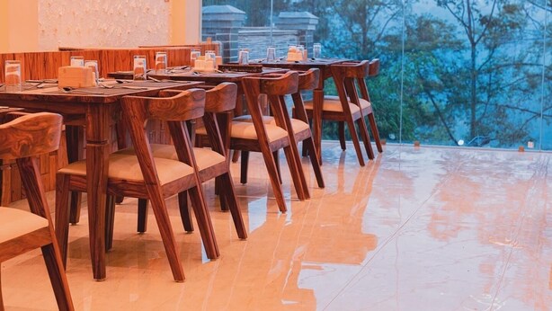 A dining area with wooden tables and chairs, a tiled floor, and a view of trees through a large window.