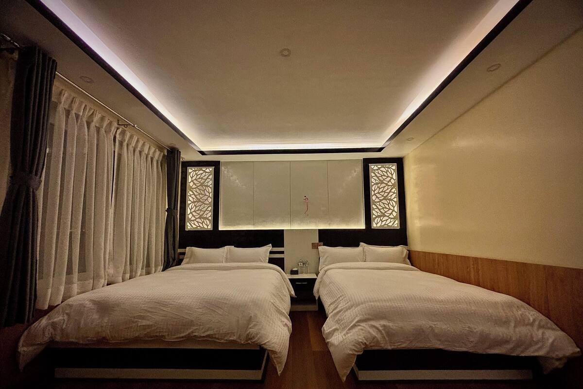 A hotel room with two beds, a nightstand, a lamp, and a decorative window blind.