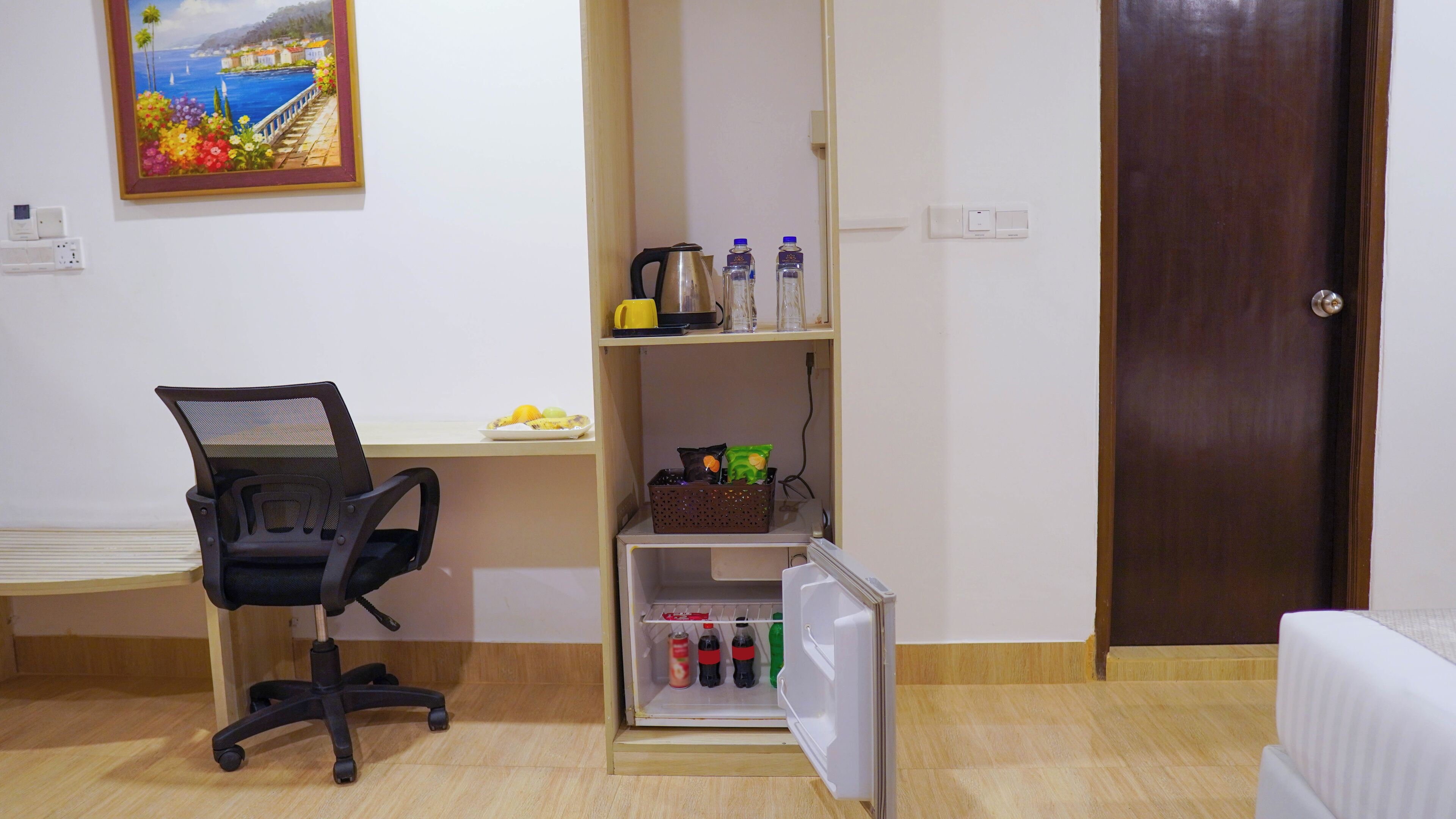 A compact hotel room with a desk, a chair, a small refrigerator, a shelf with bottles, and a door.