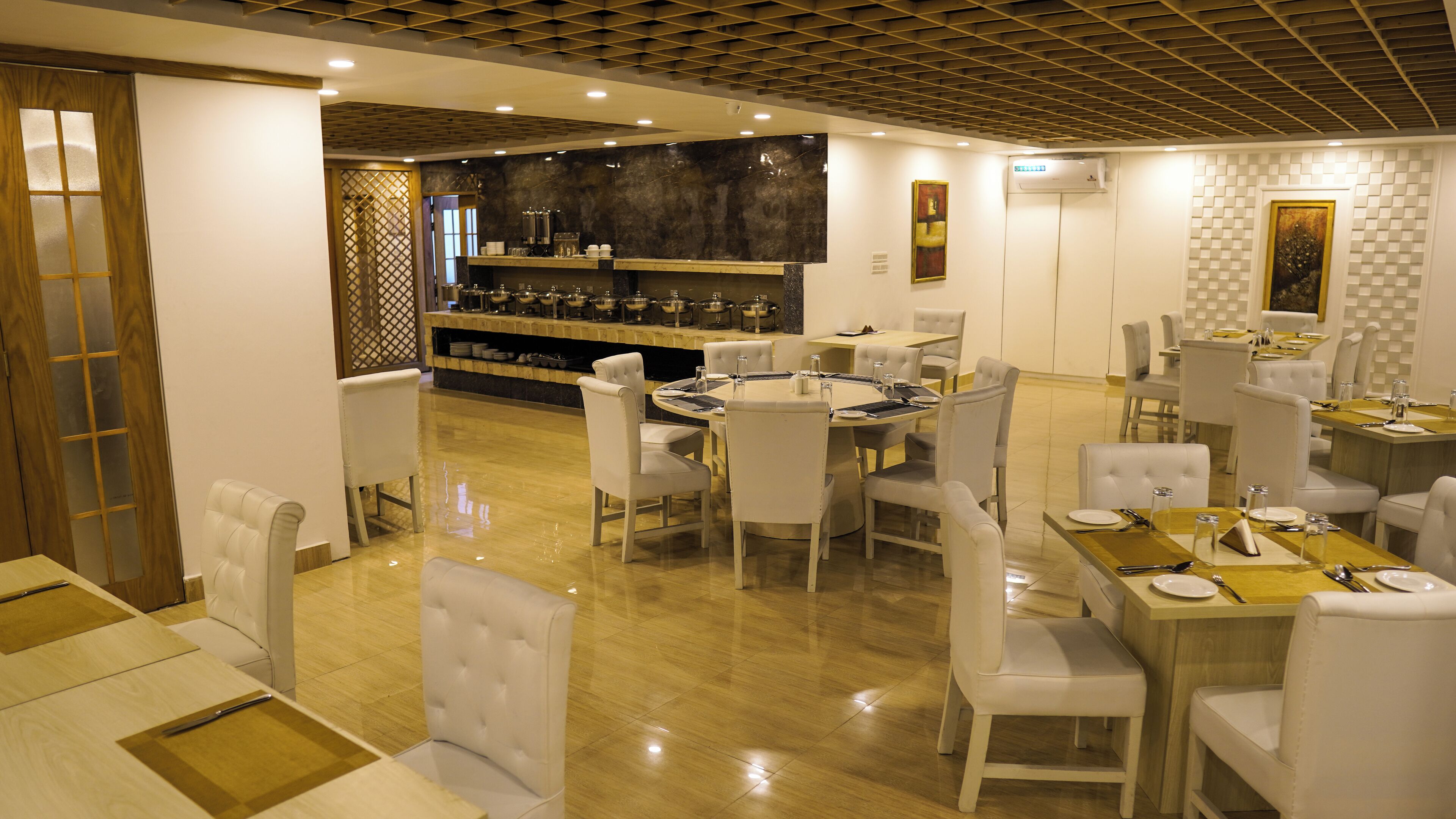 A restaurant with a central round table surrounded by white chairs, a bar area with stools, and a wooden ceiling.