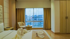 A hotel room with a bed, a chair, a small table with a plate, a window with curtains, and a wardrobe. - Grand Vistana (Dhaka)