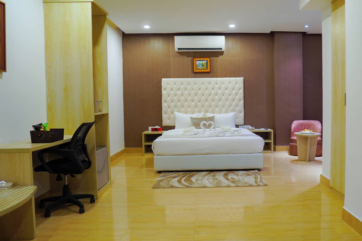A hotel room with a bed, a desk, a chair, a small table, and an air conditioning unit.