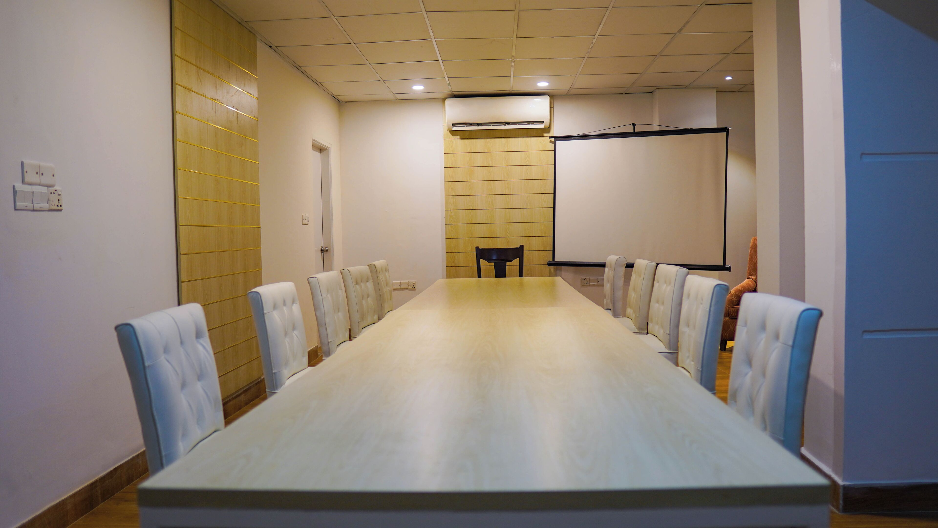 A conference room with a long table, chairs, a projector screen, and an air conditioning unit.