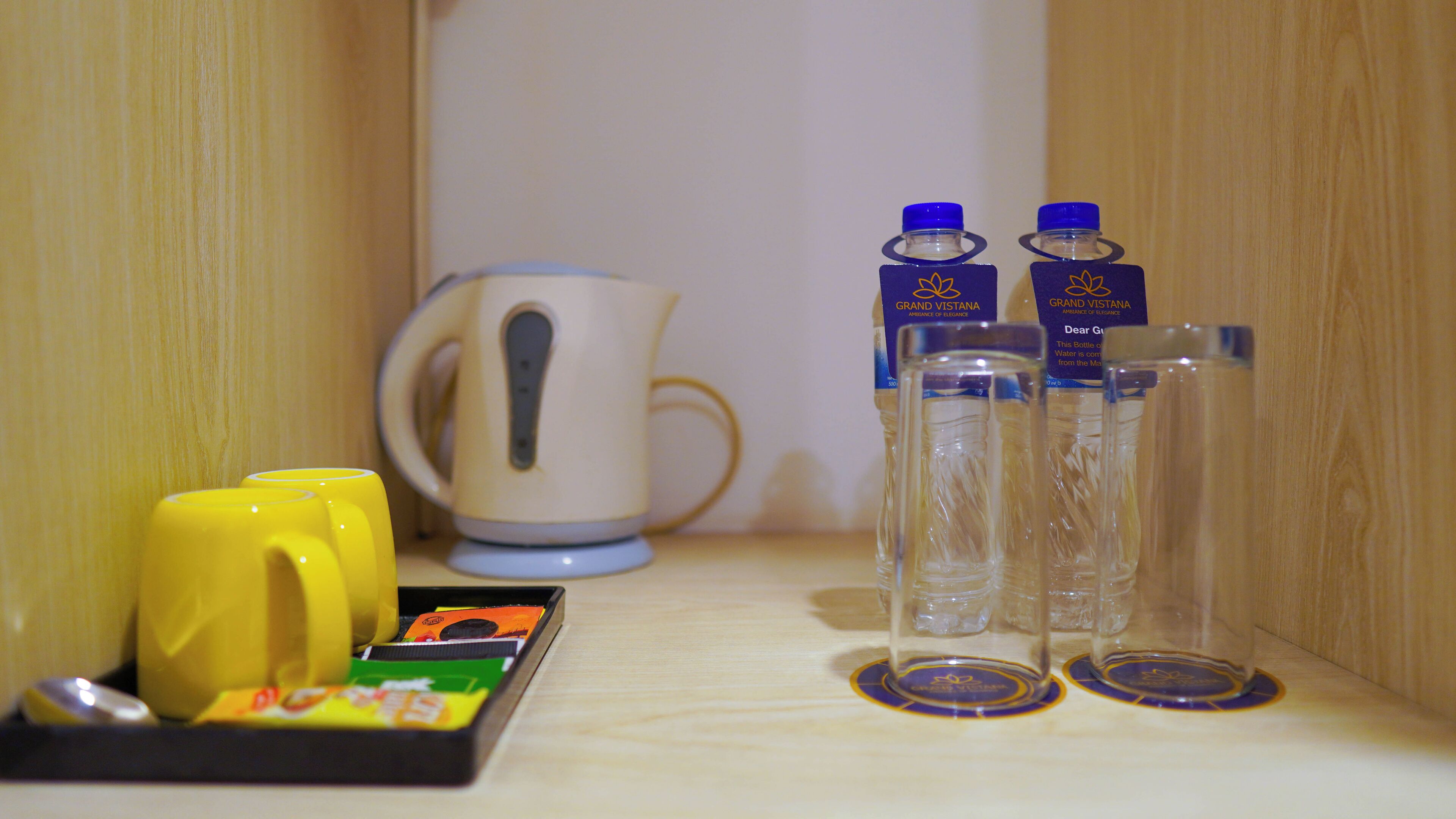 Une étagère avec une bouilloire, deux tasses jaunes, une boîte de thé et deux bouteilles d’eau.