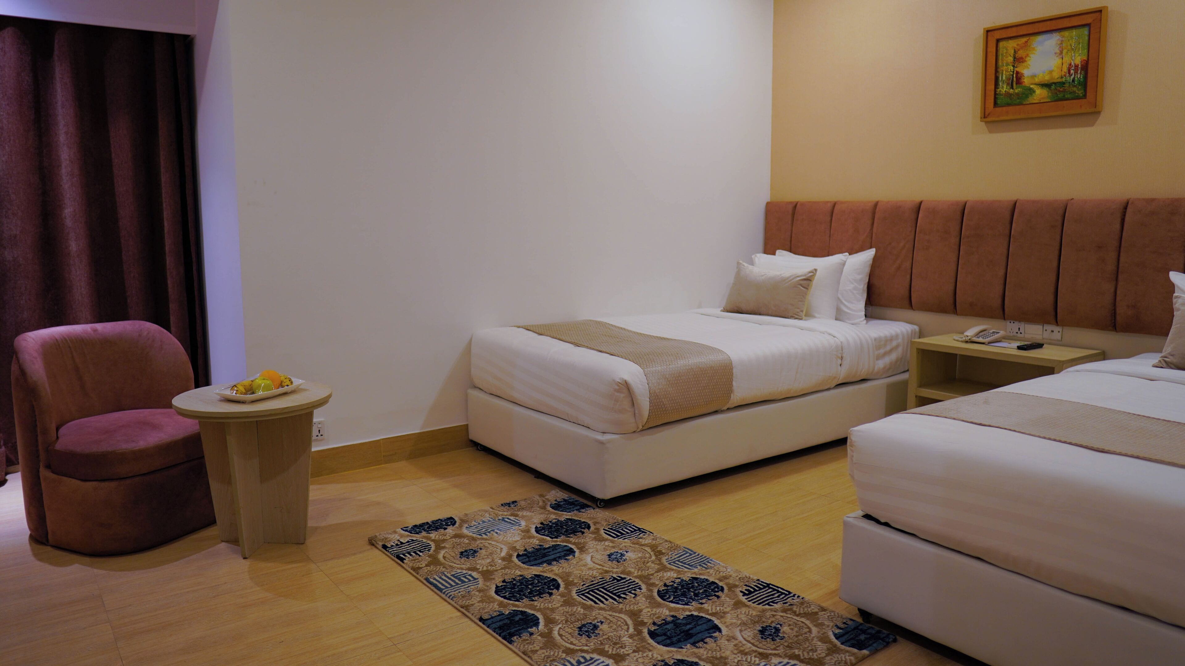 A hotel room with a bed, a chair, a small table with fruit, and a painting on the wall.