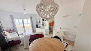 A hotel room with a wooden dining table, a chandelier, a bed with a red headboard, a balcony with a view, and a kitchenette. - Beautiful and comfortable apartment  with communal pool in Denia, Costa Blanca, Spain for 4 persons (Denia)