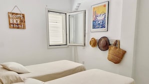 A room with two beds, a window with blinds, a 'Welcome to our BEACH HOUSE' sign, a Sydney artwork, and a straw bag. - Beautiful and comfortable apartment  with communal pool in Denia, Costa Blanca, Spain for 4 persons (Denia)
