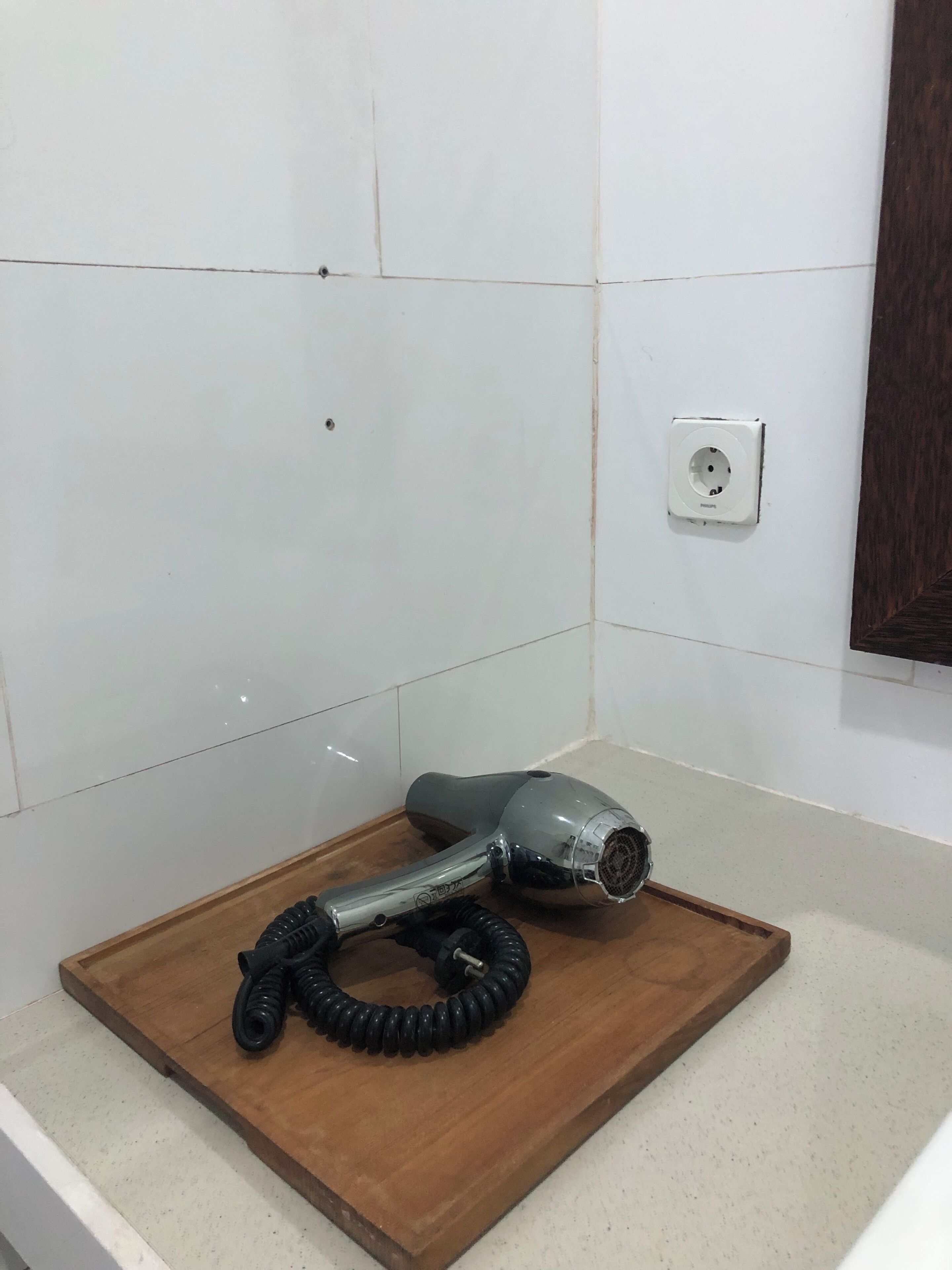 A hairdryer on a wooden tray in a bathroom with white tiles and a visible electrical outlet.