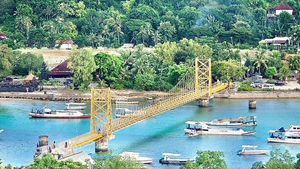 Un puente amarillo sobre un río con barcos, rodeado de un bosque verde exuberante y algunos edificios.