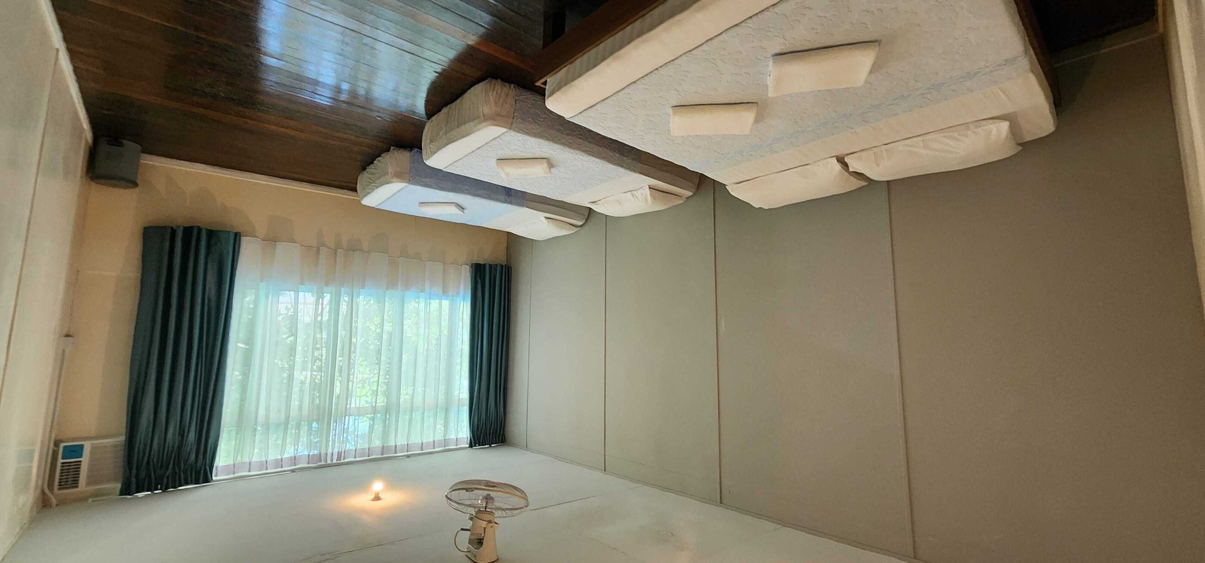 A room with a wooden ceiling, a window with sheer curtains, a standing fan, and a small table with a light.