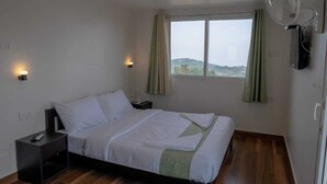 A single-bed room with a wooden floor, a window with curtains, a bedside table with a lamp, and a wall-mounted fan. - The Green Cube (Kodaikanal)