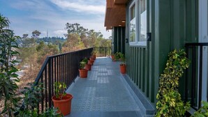 A balcony with potted plants, a metal railing, and a view of trees and buildings in the background. - The Green Cube (Kodaikanal)