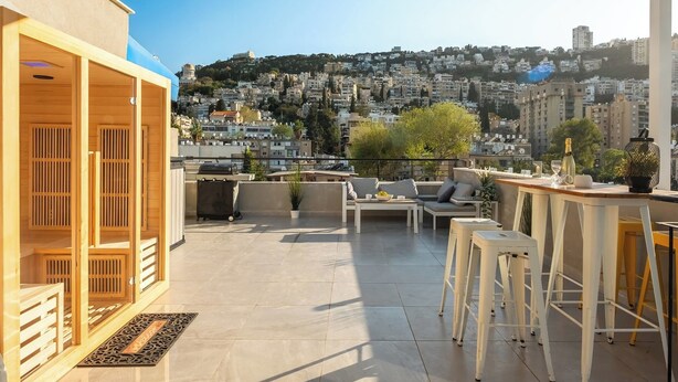 A rooftop terrace with a view of a densely built city, a grill, seating area, and a bar. - MAMAD - Bahai Gardens Penthouse by Sea N' Rent (Haifa)