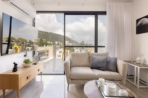 A modern living room with a sofa, a coffee table, a TV, and a large window with a city view. - MAMAD - Bahai Gardens Penthouse by Sea N' Rent (Haifa)