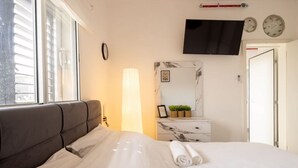 A bedroom with a bed, a bedside table with a lamp, a TV mounted on the wall, and a door. - MAMAD - Bahai Gardens Penthouse by Sea N' Rent (Haifa)