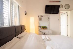 Un dormitorio con cama, lámpara de noche, televisor montado en la pared, reloj y puerta.
