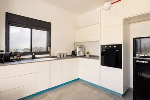 A modern kitchen with white cabinets, a black refrigerator, a coffee maker, and a window with a city view. - MAMAD - Bahai Gardens Penthouse by Sea N' Rent (Haifa)