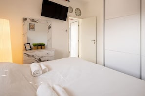 Quarto de hotel com cama, televisão fixada na parede, mesa de cabeceira com abajur, espelho e porta.