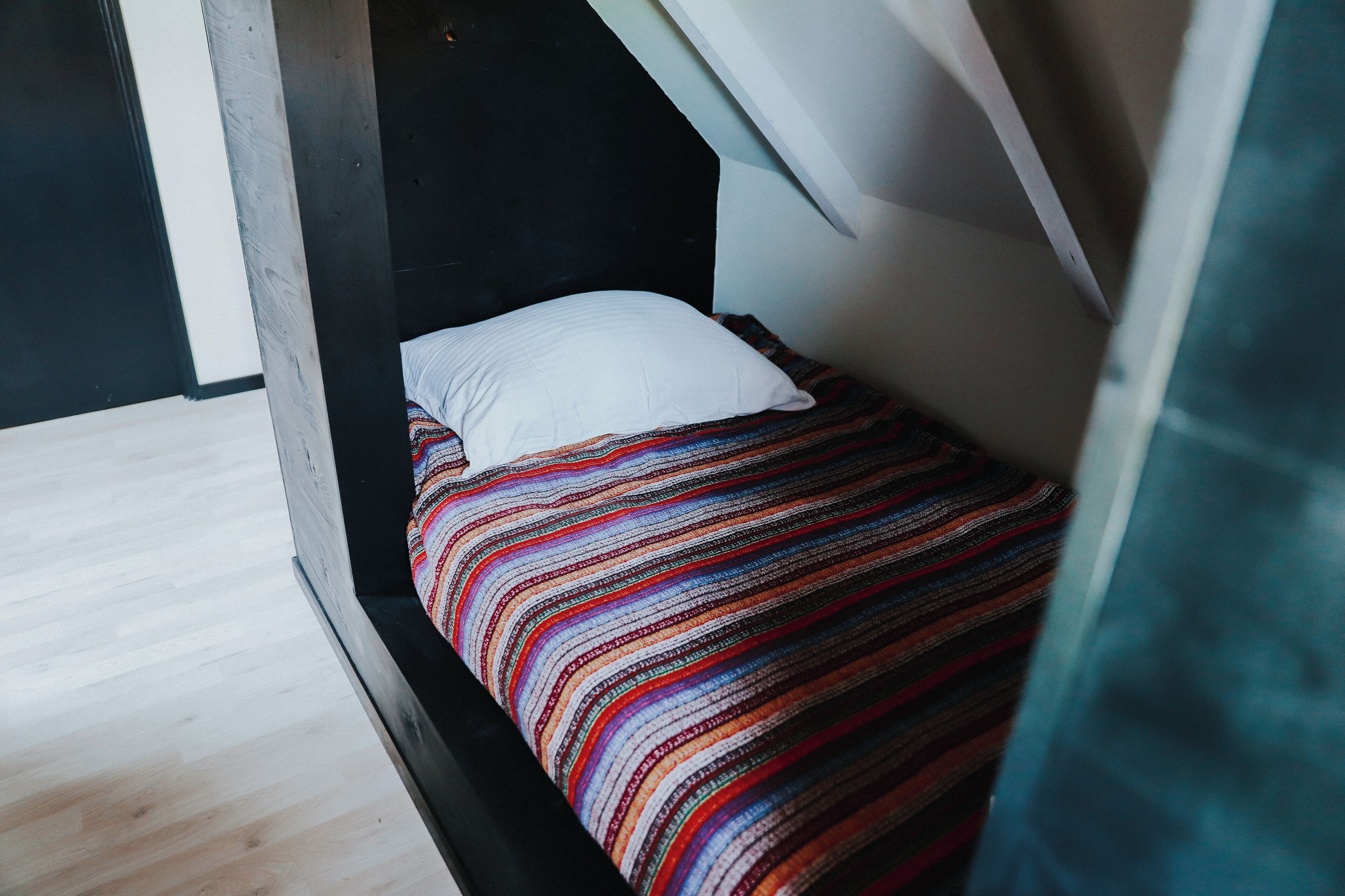 Een kleine, smalle kamer met een schuine balkenlaag, een enkel bed met een kleurrijke gestreepte deken en een wit kussen.