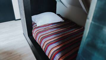Ein kleiner, schmaler Raum mit schräger Decke, einem Einzelbett mit farbenfroher Streifenoptik und einem weißen Kopfkissen.