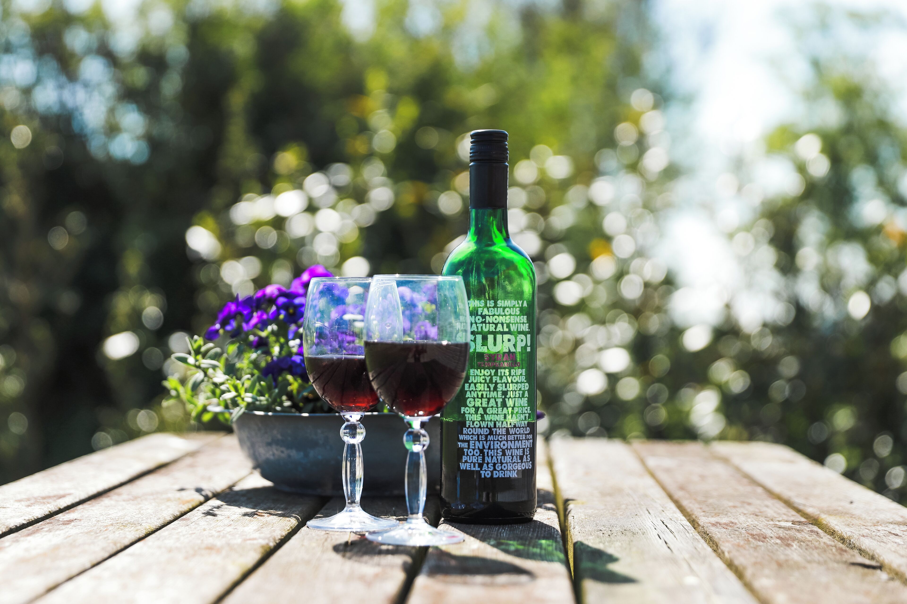 Una botella de vino y dos copas sobre una mesa de madera, con un jardín desenfocado al fondo.