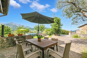 Eine Terrasse im Freien mit einem Holztisch, Korbstühlen, einem grünen Sonnenschirm und Blumentöpfen.