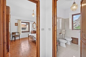 Ein Zimmer mit einem Bett, einem Sessel, einem Fenster mit Vorhängen sowie einem Badezimmer mit Toilette und Bidet.