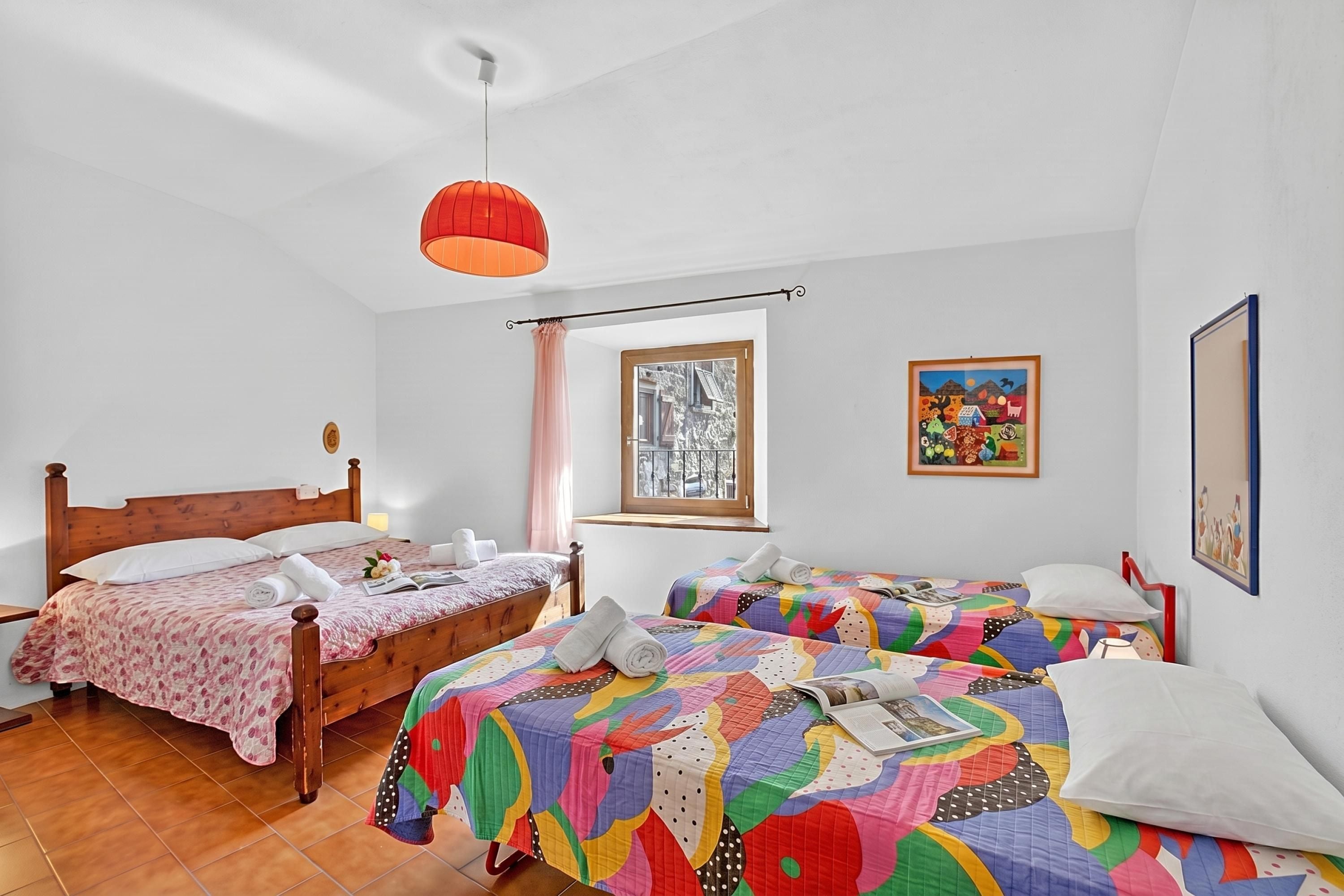 A room with a wooden bed, a colorful quilted bedspread, a red pendant light, and two paintings on the wall.
