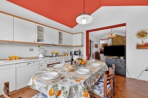 Uma cozinha moderna com armários brancos, uma mesa de jantar posta para quatro pessoas e uma televisão ao fundo.