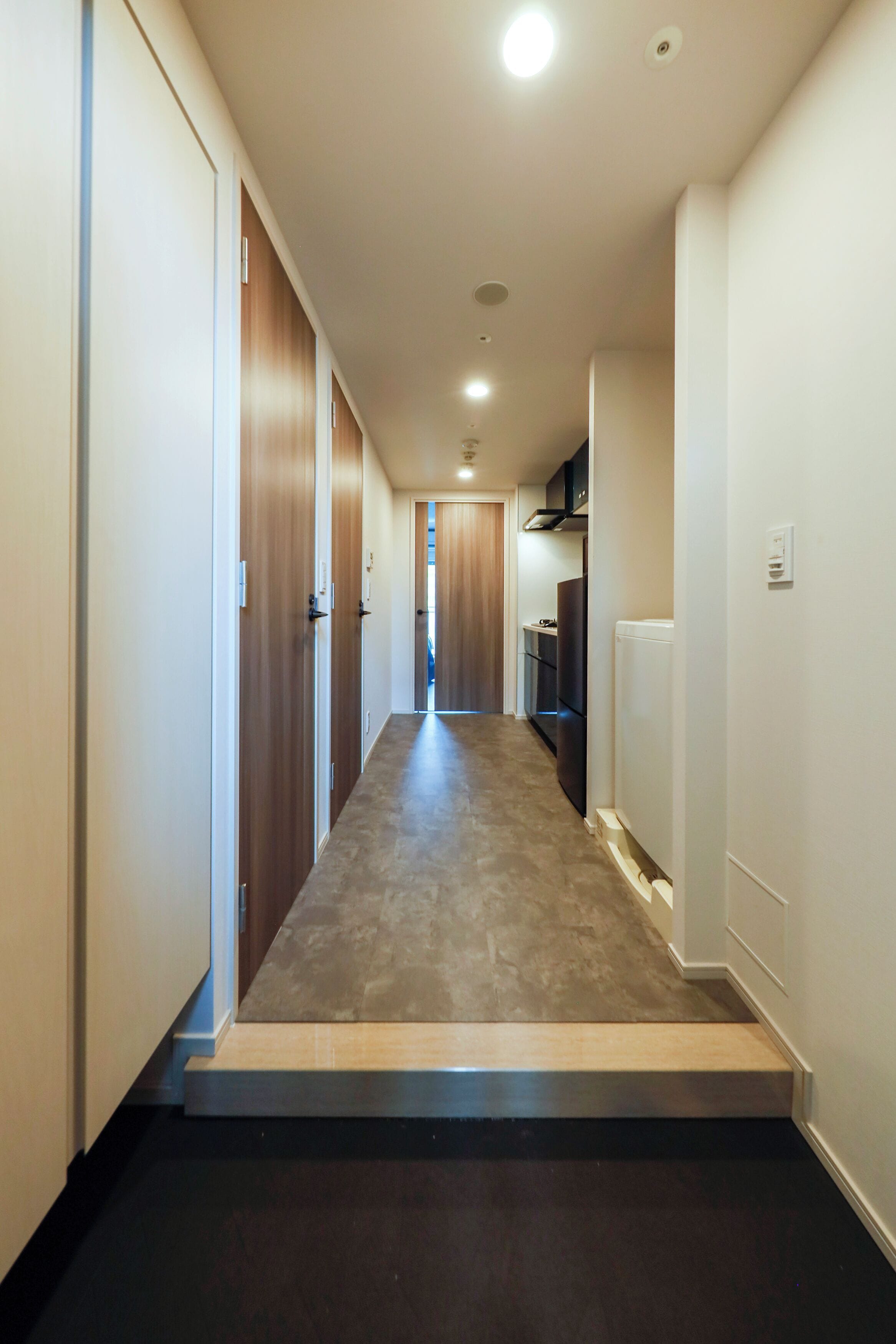 A narrow hallway with wooden doors on one side and a kitchen area on the other.