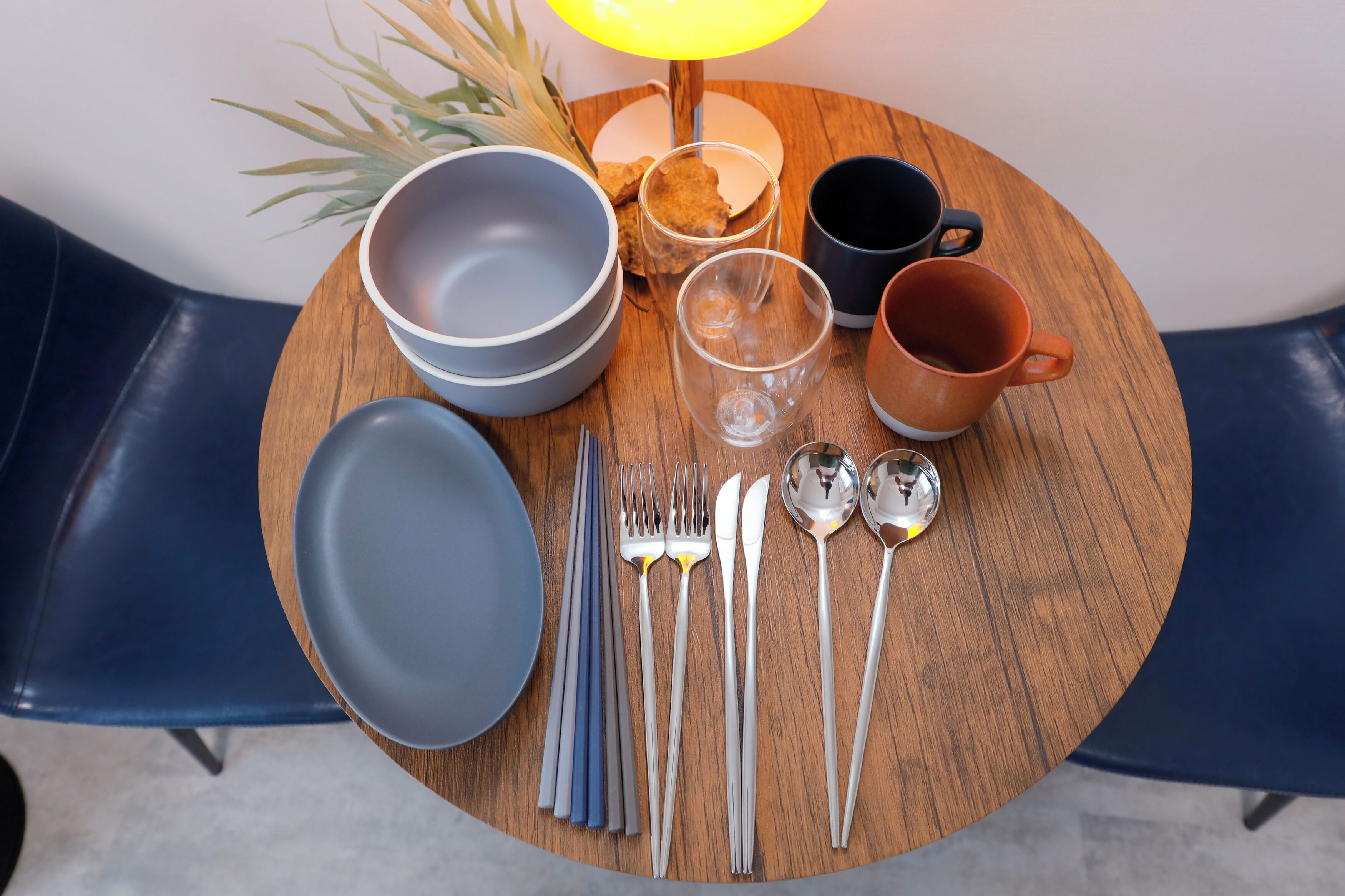 A round wooden table with a set of cutlery, bowls, glasses, and a lamp.