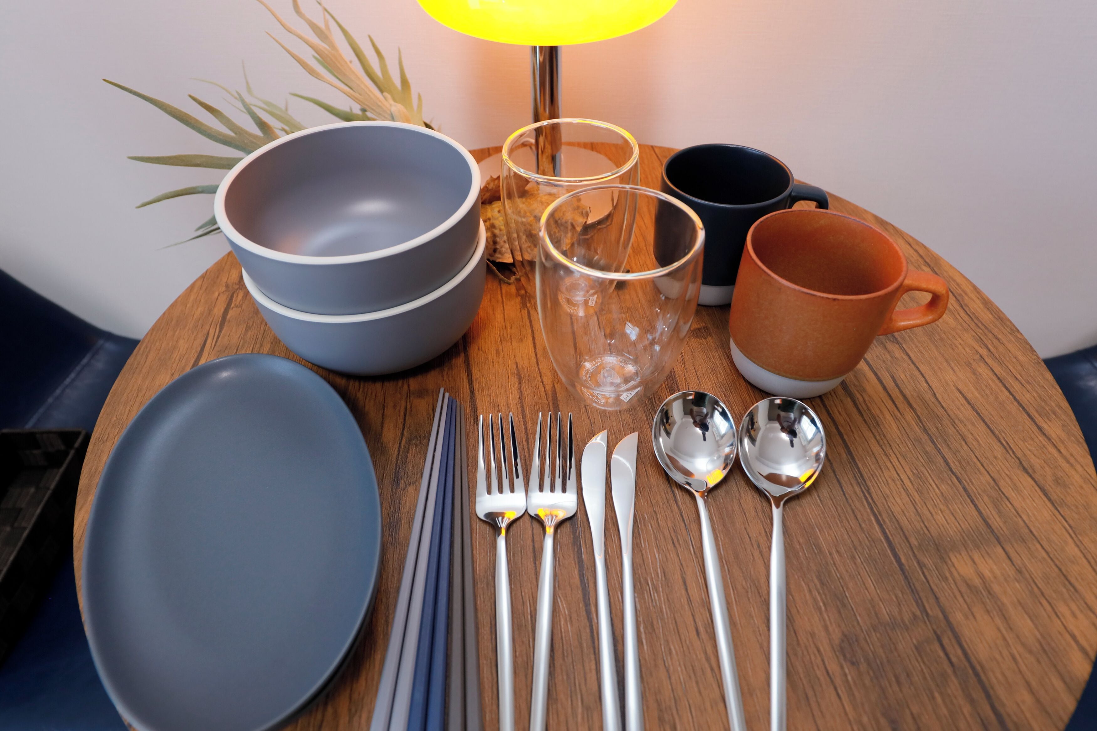 A wooden table with a set of blue and gray plates, clear glasses, a brown mug, and silver cutlery.