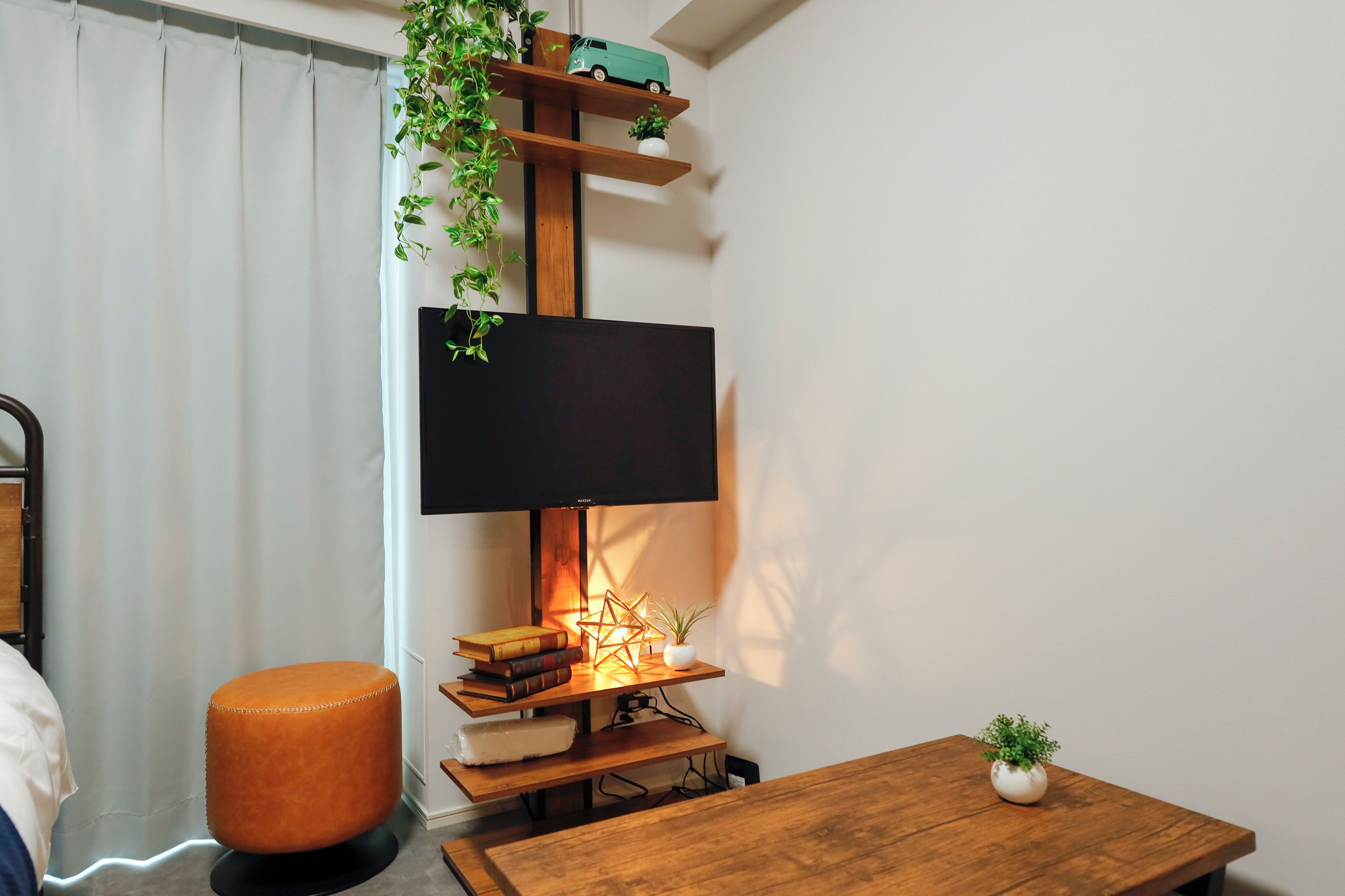 A modern living room with a flat-screen TV mounted on a wooden stand, a small table with a plant, and a cushioned stool.