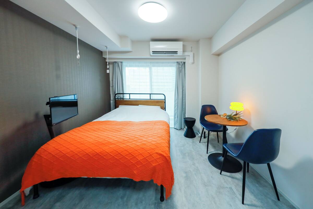 Habitación de hotel con cama, escritorio, silla, una mesita con lámpara y equipo de aire acondicionado.