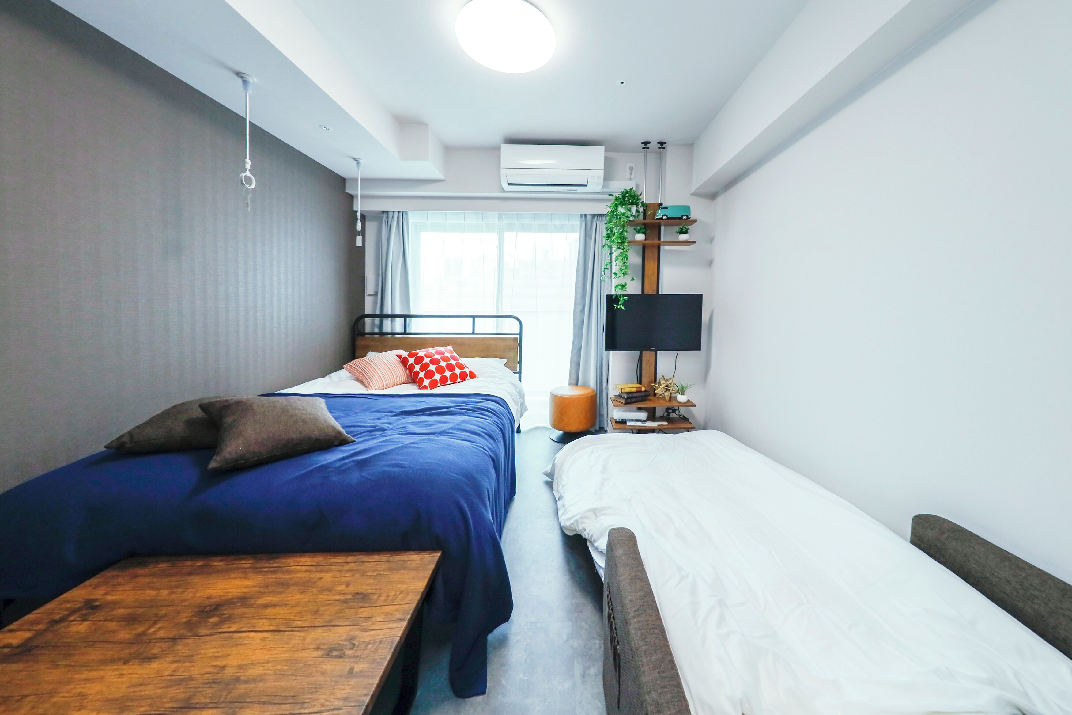 A compact hotel room with two beds, a wooden table, a TV, and a small shelf with decorative items.