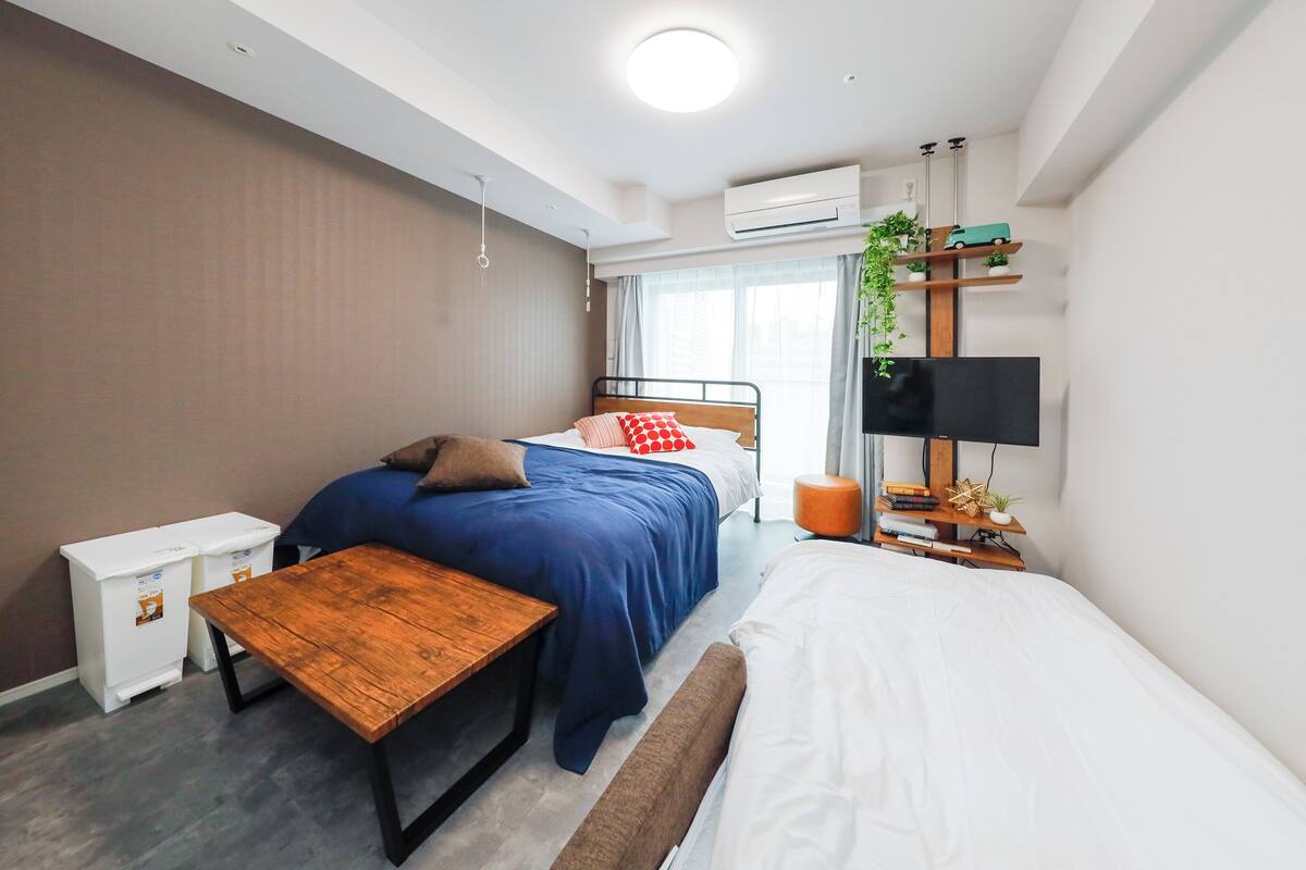 Un dormitorio moderno con cama, mesita de noche y un pequeño banco de madera. Hay un televisor en un mueble, un estante con objetos decorativos y un equipo de aire acondicionado en la pared.